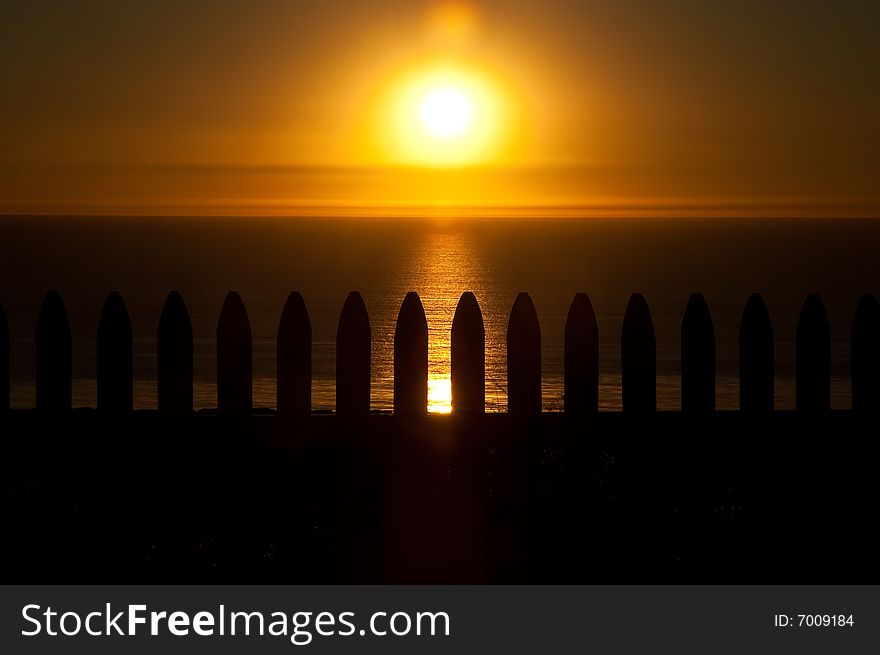 The sun sets on the Pacific Ocean over a white picket fence, silhouetted by the sun. The sun sets on the Pacific Ocean over a white picket fence, silhouetted by the sun.