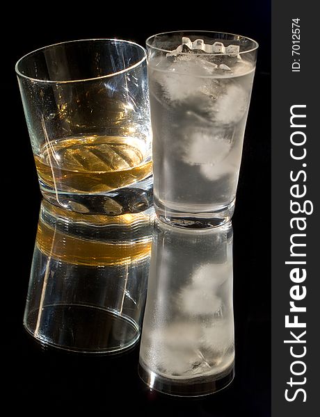 A glass of whiskey and a glass of ice water against a black background, reflected in the surface. A glass of whiskey and a glass of ice water against a black background, reflected in the surface.
