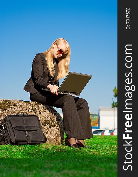 Blond businesswoman in sunglasses working on laptop sitting on a stone in park. Blond businesswoman in sunglasses working on laptop sitting on a stone in park