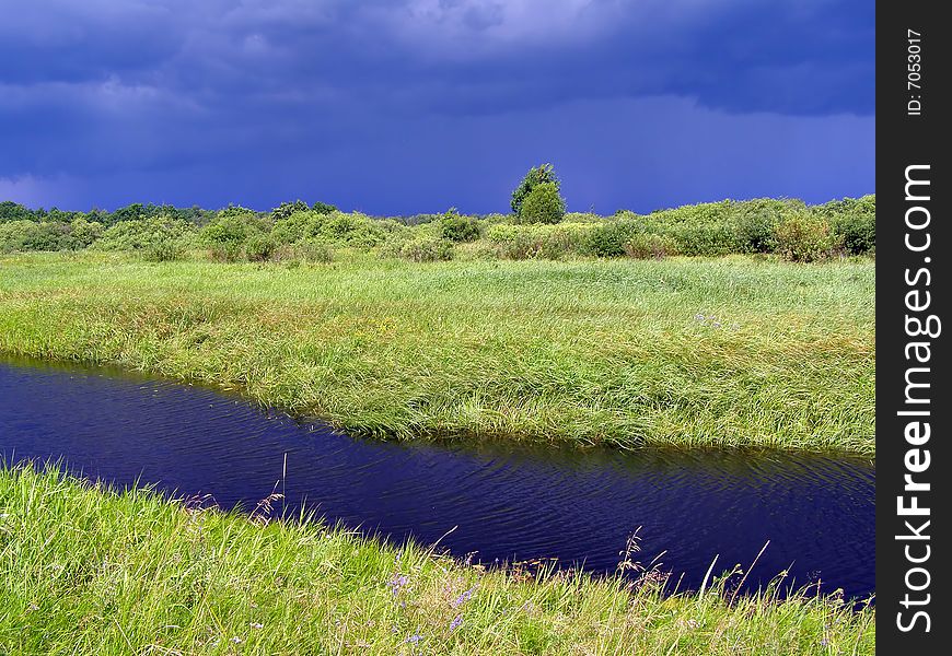 Small river on green field before thunderstorm