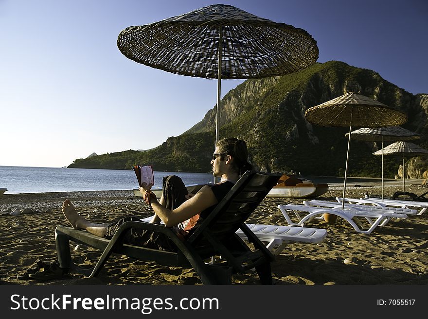 Young man sittin on the beach under umbrella reading a book. Young man sittin on the beach under umbrella reading a book