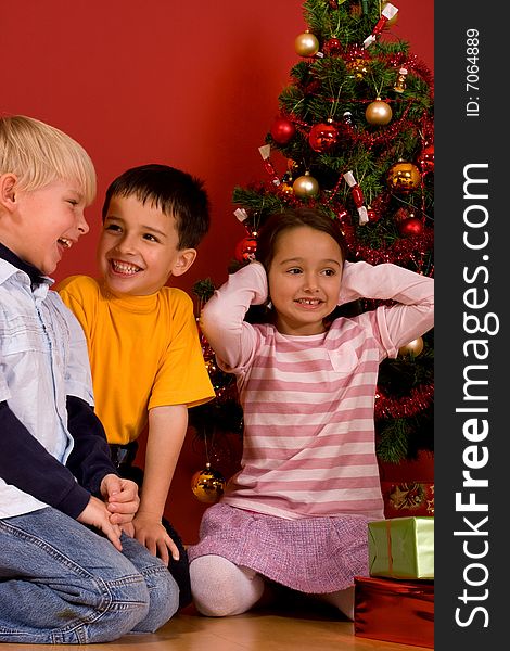 Smiling children sitting by Christmas tree in red room