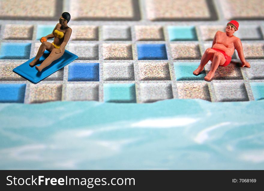 A darker-skinned woman is sunbathing near a sunburned overweight man. They are on a tile patio by a pool. They are miniature figures. Copyspace at bottom. A darker-skinned woman is sunbathing near a sunburned overweight man. They are on a tile patio by a pool. They are miniature figures. Copyspace at bottom.