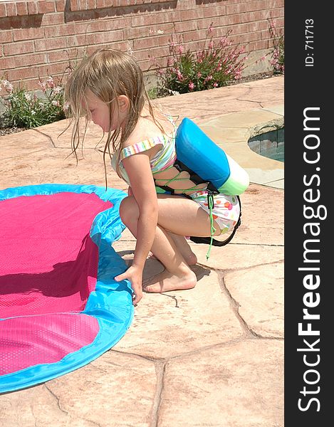 Toddler wears her floating vest to keep her safe if she falls in the pool. She is picking up a float that she needs aired up. Toddler wears her floating vest to keep her safe if she falls in the pool. She is picking up a float that she needs aired up.