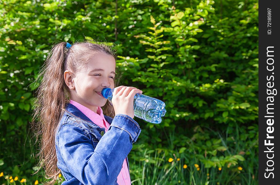 Girl in a blue denim jacket, drinks water from a plastic bottle outside. Girl in a blue denim jacket, drinks water from a plastic bottle outside