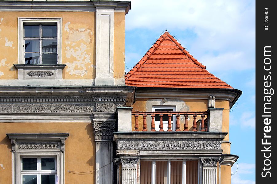 Fragment of a classic building, with pretty balcony with rail, and red roof. Peeling yellow paint and blue sky. Sculpted frieze. Fragment of a classic building, with pretty balcony with rail, and red roof. Peeling yellow paint and blue sky. Sculpted frieze.