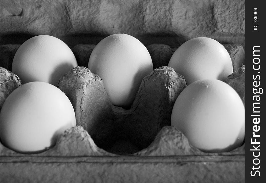 Five eggs in soft, dim light in a grey carton, arranged as to imply that a central egg has been removed. Grescale image.