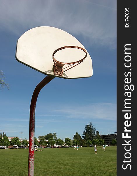 Rural basketball hoop without net and fields with playing kids in background.