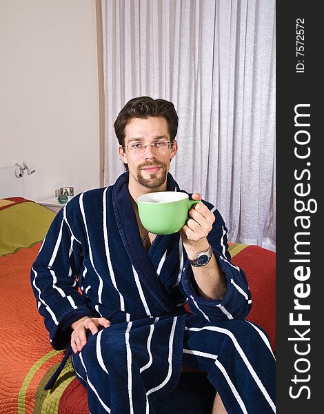 Young man in glasses wearing blue striped bathrobe sitting in his bedroom and drinking coffee or tea from green cup. Young man in glasses wearing blue striped bathrobe sitting in his bedroom and drinking coffee or tea from green cup