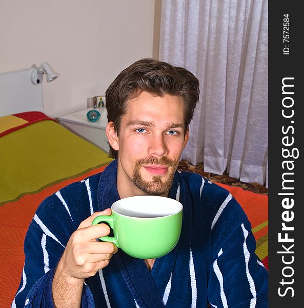 Young man in glasses wearing blue striped bathrobe sitting in his bedroom and drinking coffee or tea from green cup. Young man in glasses wearing blue striped bathrobe sitting in his bedroom and drinking coffee or tea from green cup