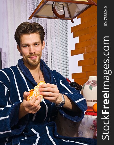 Young man wearing blue striped bathrobe sitting in his kitchen and peeling tangerine. Young man wearing blue striped bathrobe sitting in his kitchen and peeling tangerine