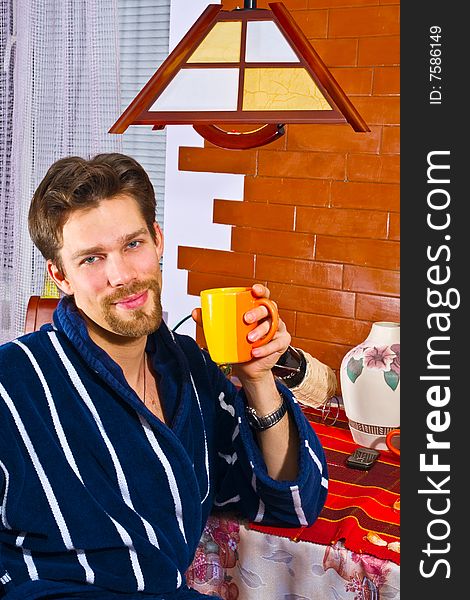 Young man wearing blue striped bathrobe sitting in his kitchen and drinking his breakfast tea. Young man wearing blue striped bathrobe sitting in his kitchen and drinking his breakfast tea