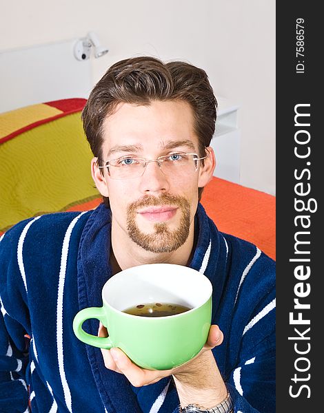 Young man in glasses wearing blue striped bathrobe sitting in his bedroom and drinking coffee or tea from big green cup. Young man in glasses wearing blue striped bathrobe sitting in his bedroom and drinking coffee or tea from big green cup