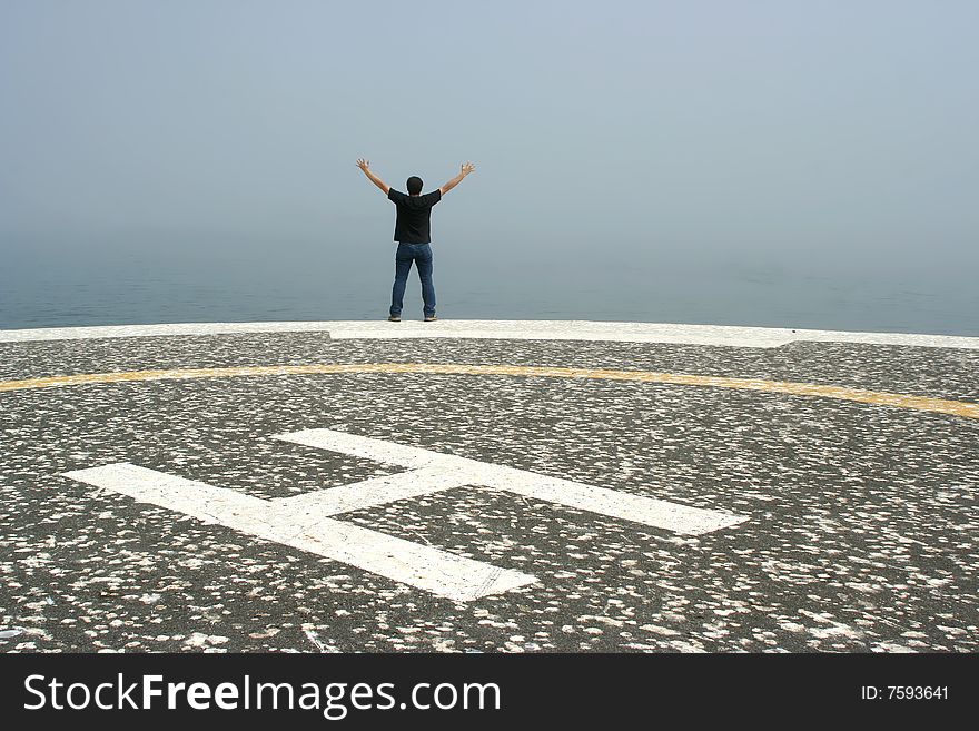 Young man with arms open contemplating the ocean in foggy day in the heliport - freedom or success concept