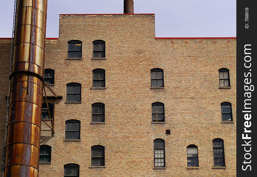 Windows and a rusted smokestack against a brown or tan brick wall on the side of an urban apartment building.