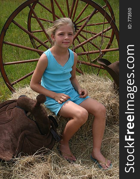 Girl sitting ontop of hay next to a saddle, wheel and hat. Girl sitting ontop of hay next to a saddle, wheel and hat