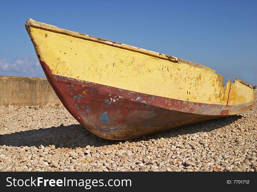 A battered boat or skiff resting on a gravel beach under a blue sky