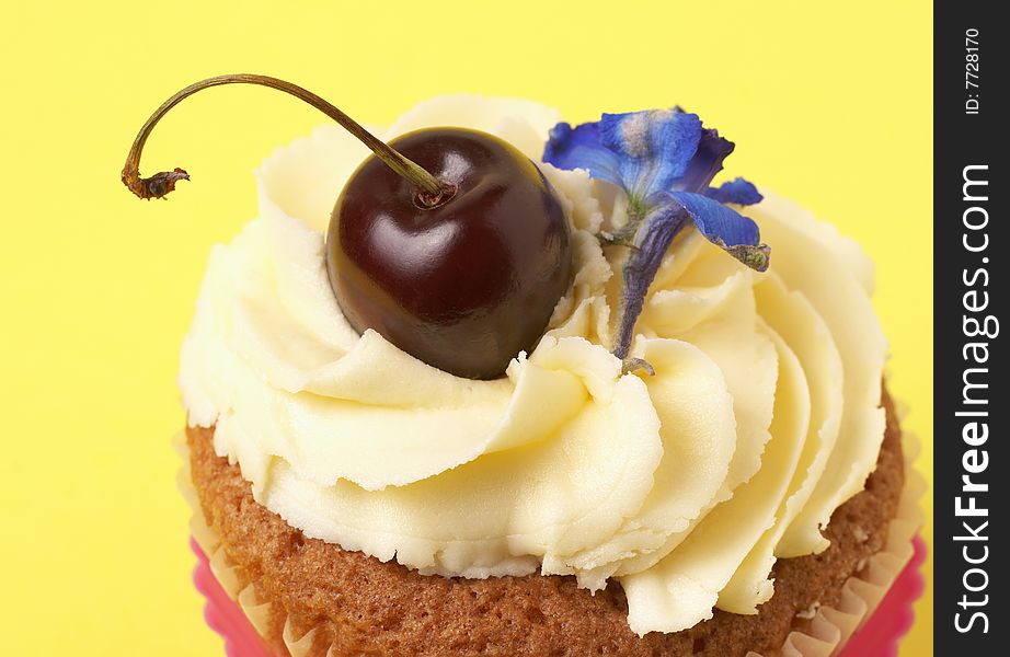Miniature vanilla cupcake with icing, fresh cherry and blue flower on yellow background. Miniature vanilla cupcake with icing, fresh cherry and blue flower on yellow background