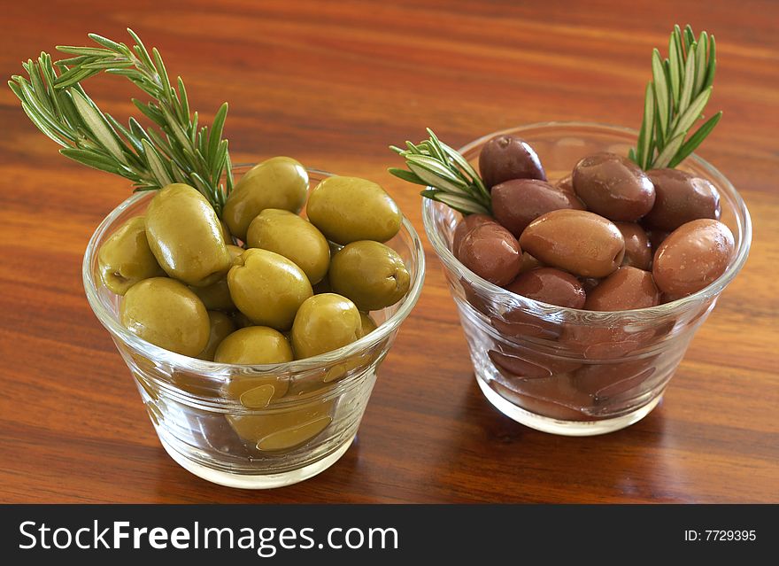 Two jars of green and black olives with stick of rosemary on wooden table background