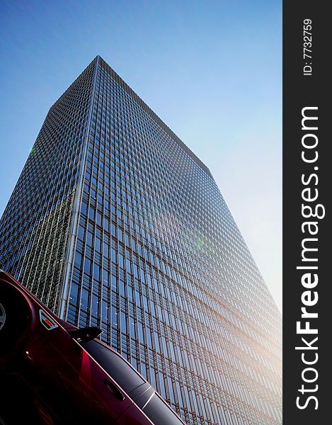 Part of car parked at angle with tall office building in background reaching up to the sky with sun glare on faÃ§ade. Part of car parked at angle with tall office building in background reaching up to the sky with sun glare on faÃ§ade.