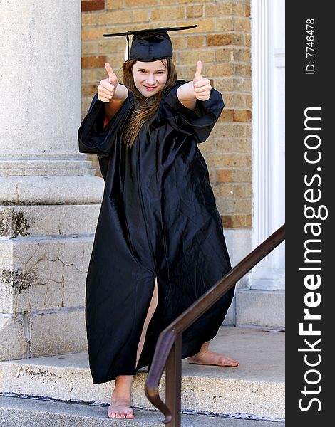 A barefoot middle school graduate in cap and gown, displaying thumbs up on the steps of her school. A barefoot middle school graduate in cap and gown, displaying thumbs up on the steps of her school.