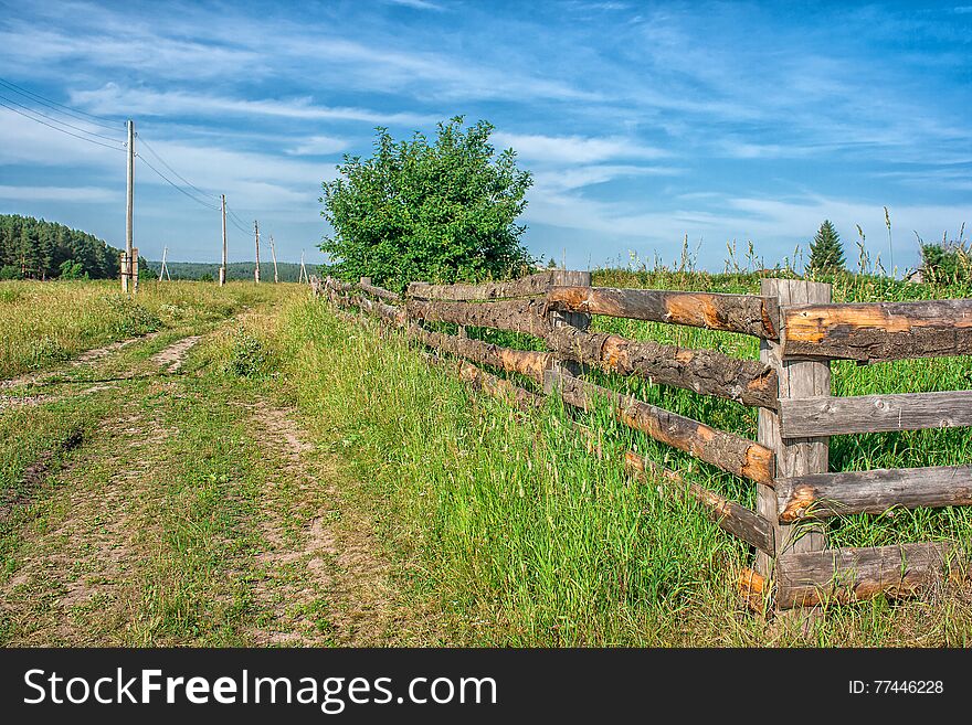 Rural landscape with wooden fence, road, meadow and white clouds on blue sky