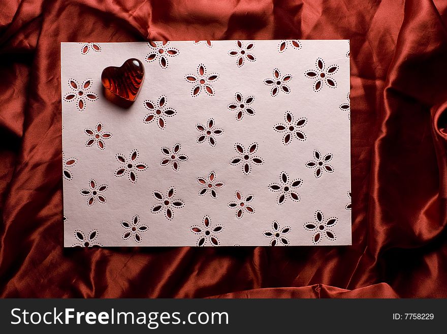 A pink sheet of paper decorated with flowers and a red glass heart, lying on red satin background. A pink sheet of paper decorated with flowers and a red glass heart, lying on red satin background.