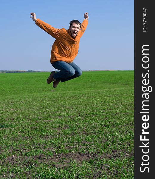 Jumping man in jeans and orange jacket on blue sky and green grass background