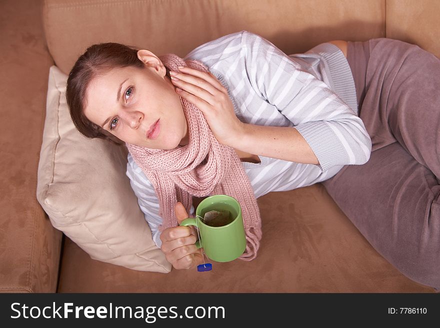 A young woman is sick. She is lying on the couch and is sneezing. She has a tea in her hand. A young woman is sick. She is lying on the couch and is sneezing. She has a tea in her hand.