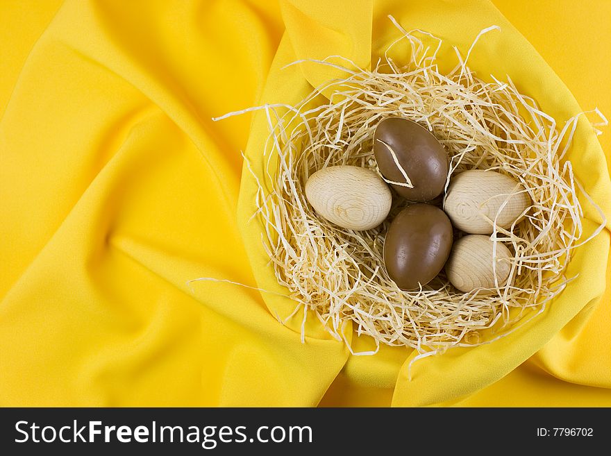 Wooden and chocolate easter eggs placed in a decorative nest, yellow fabric background.