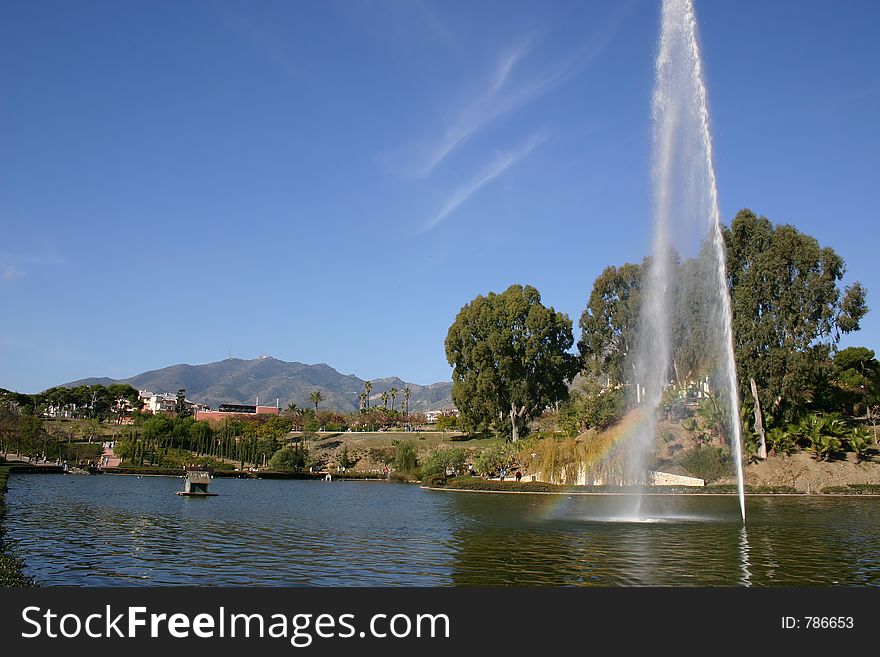 There is a rainbow in the fountain on this lovely lake scene. There is a rainbow in the fountain on this lovely lake scene