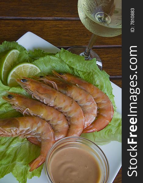 Fresh king prawns placed on a bed of lettuce served with a pink sauce and slices of lemons accompanied by a glass of white wine in an outdoor setting.