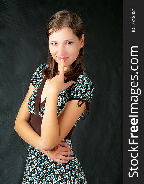 Young caucasian girl standing half-turned and wearing colored dress with brown belt,metal chaing bracelet,smiling with hands crossed in front,isolated on black