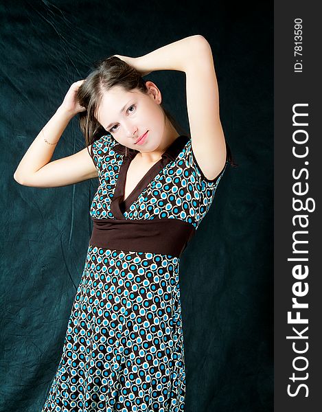 Young caucasian girl with soft skin standing turned with hands up crossed behind head standing tilted looking left side wearing colored dress with brown belt isolated on black. Young caucasian girl with soft skin standing turned with hands up crossed behind head standing tilted looking left side wearing colored dress with brown belt isolated on black