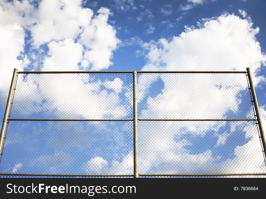 A big blue sky with many clouds stands above a gated linked fence outside in the summer time. A big blue sky with many clouds stands above a gated linked fence outside in the summer time