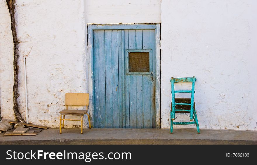 Two old chairs standing on a sidwalk in front of a wall with a blue door. Two old chairs standing on a sidwalk in front of a wall with a blue door