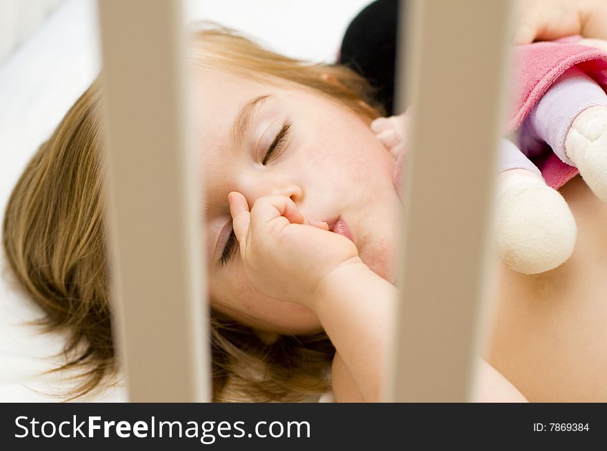 Beautiful baby girl sucking her thumb and hugging her doll in her cot just before bedtime