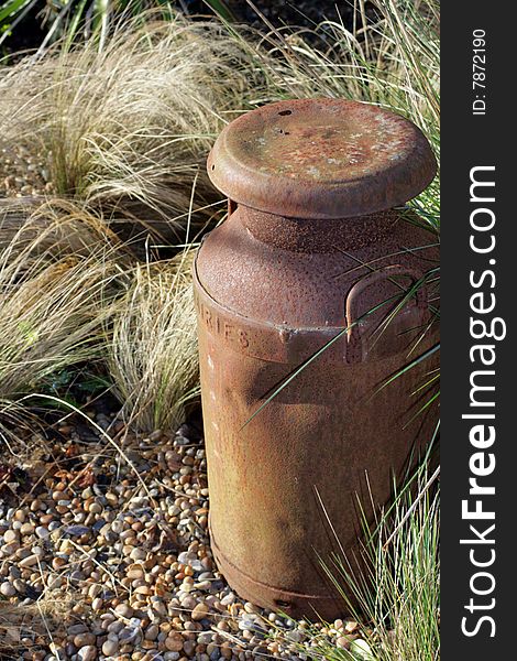 A rusty milk churn set amongst dry grasses on a pebbled path base. Used as a garden ornament.