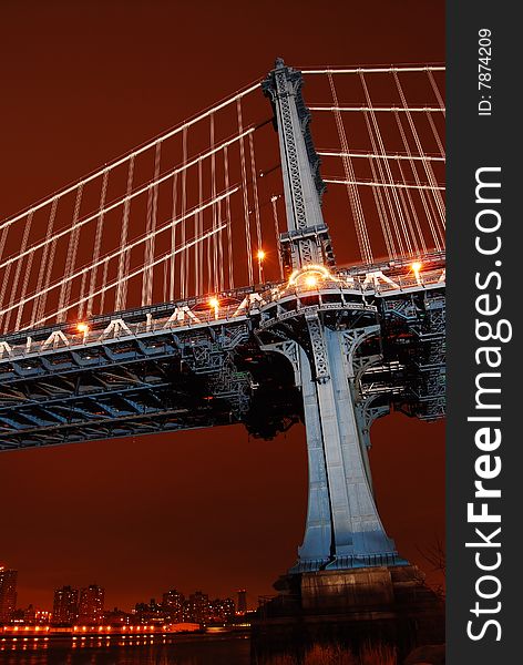 This is a shot of one of the vertical support structures of the Manhattan Bridge. The shot was taken just after sundown. The location was on the Brooklyn side of the Bridge. The location is between the Brooklyn Bridge and looking up from the park area below. The bridge is lit by floodlights provided by the city and appears light blue in the image. The sky has a dark reddish brown tone in the color. A very small portion of Manhattan can be seen in the lower part of the image, by the river. This is a shot of one of the vertical support structures of the Manhattan Bridge. The shot was taken just after sundown. The location was on the Brooklyn side of the Bridge. The location is between the Brooklyn Bridge and looking up from the park area below. The bridge is lit by floodlights provided by the city and appears light blue in the image. The sky has a dark reddish brown tone in the color. A very small portion of Manhattan can be seen in the lower part of the image, by the river.
