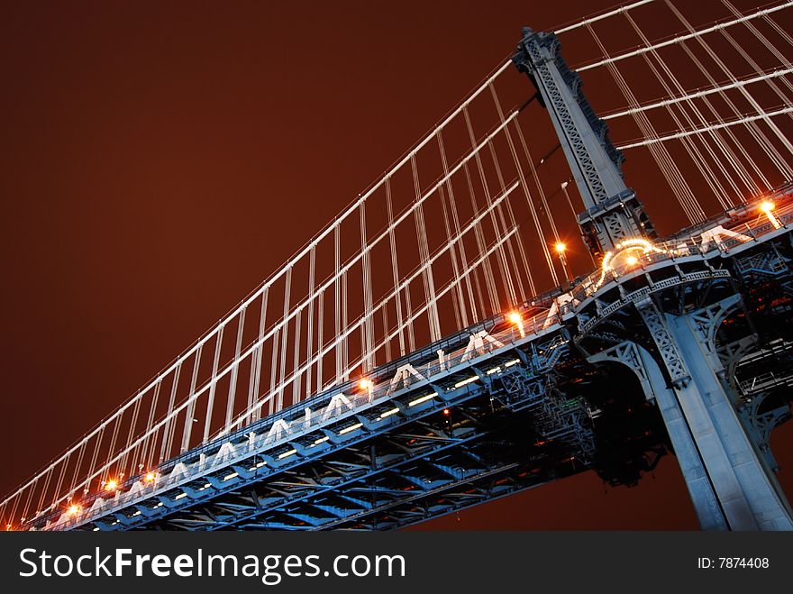 This is a shot of one of the vertical support structures of the Manhattan Bridge. The shot was taken just after sundown. The location was on the Brooklyn side of the Bridge. The location is between the Brooklyn Bridge and looking up from the park area below. The bridge is lit by floodlights provided by the city and appears light blue in the image. The sky has a dark reddish brown tone in the color. Neither the Hudson river or the city of Manhattan are visible in this image. This is a shot of one of the vertical support structures of the Manhattan Bridge. The shot was taken just after sundown. The location was on the Brooklyn side of the Bridge. The location is between the Brooklyn Bridge and looking up from the park area below. The bridge is lit by floodlights provided by the city and appears light blue in the image. The sky has a dark reddish brown tone in the color. Neither the Hudson river or the city of Manhattan are visible in this image.