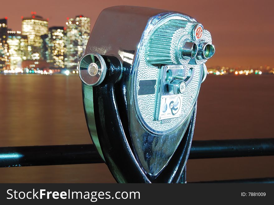 This is a shot of a coin operated viewfinder. This viewfinder is found on the Jersey side of the Hudson river. In the background the city of Manhattan can be seen. The subject is not the city but the finder itself. The image was captured at night. Lighting was provided by some local street lights. The finder is ever slightly tilted back. It is rotated on its axis so the back and part of one side is seen. A very small section on top of the finder is seen. The color of the finder is silver. The background has a brownish look and shows the city and part of the river in the image. A protective fall off rail can be seen running the width of the image in the lower third. This is a shot of a coin operated viewfinder. This viewfinder is found on the Jersey side of the Hudson river. In the background the city of Manhattan can be seen. The subject is not the city but the finder itself. The image was captured at night. Lighting was provided by some local street lights. The finder is ever slightly tilted back. It is rotated on its axis so the back and part of one side is seen. A very small section on top of the finder is seen. The color of the finder is silver. The background has a brownish look and shows the city and part of the river in the image. A protective fall off rail can be seen running the width of the image in the lower third.