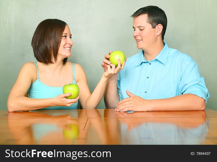 Beautiful young female caucasian offering green apple to her boyfriend or husband on kitchen table. Beautiful young female caucasian offering green apple to her boyfriend or husband on kitchen table