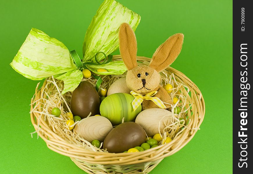 A toy easter bunny in a wicker basket with eggs and ribbon in green and yellow.