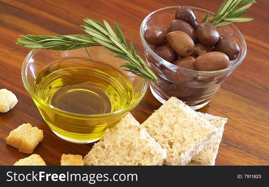 Two jars of black olives with stick of rosemary, olive oil, slices of bread and croutons on wooden table background. Two jars of black olives with stick of rosemary, olive oil, slices of bread and croutons on wooden table background