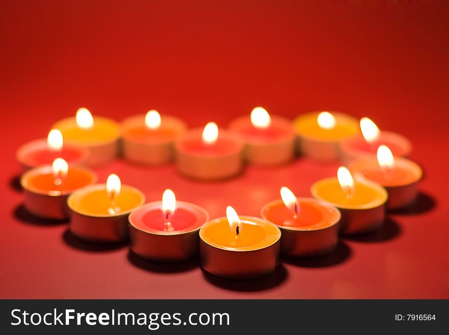 Small light candles, in heart form on a red background. Focus on the first, front candle. Small light candles, in heart form on a red background. Focus on the first, front candle.