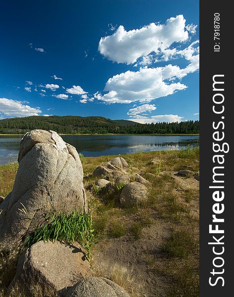 A landscape with weathered boulders sitting in front of a pristine lake and green, wooded mountains in the background. A landscape with weathered boulders sitting in front of a pristine lake and green, wooded mountains in the background