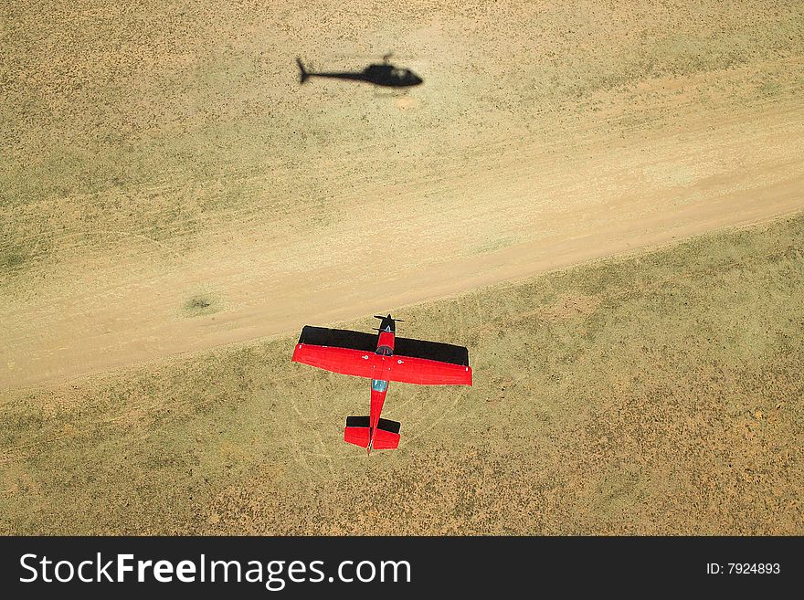 Aerial photograph of a red Cessna airplane next to a dirt airstrip with shadow of a helicopter