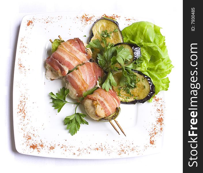 Grilled chiken meat wraped in bacon stringed on wooden brochette decorated with grilled egg-plant, leaves of salad and parsley on white plate