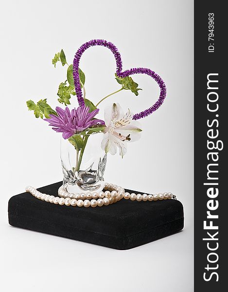 Valentine's day still life: a flower arrangement in a glass with a purple heart and a string of pearls all on a black jewelry box with a white background. Valentine's day still life: a flower arrangement in a glass with a purple heart and a string of pearls all on a black jewelry box with a white background