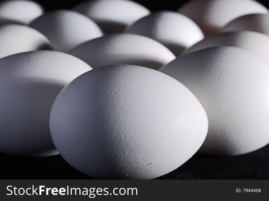A lots of white chicken eggs on black background, close-up. Focus is on the egg at the front. A lots of white chicken eggs on black background, close-up. Focus is on the egg at the front.